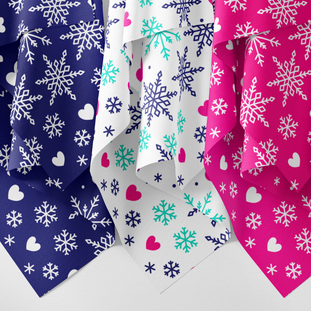 Image of three hanging pieces of fabric with a fairisle snowflake design. One in blue with a white design, one in white with pink, aqua and blue design and one in pink with a white design