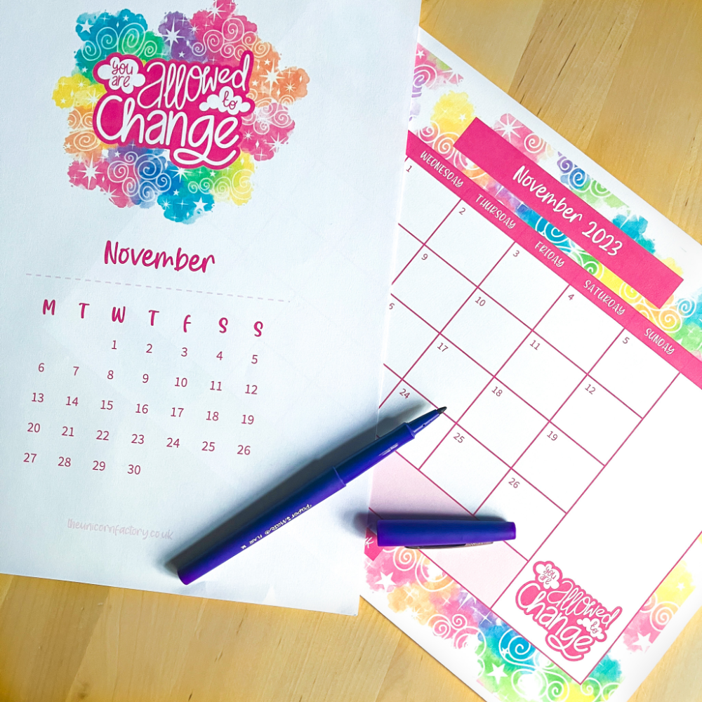 A photograph of two printed November 2023 calendars on a table. The calendar on the left has the illustrated quote "you are allowed to change" surrounded by clouds and stars in rainbow colours above a grid of November dates. On the right is a November planner calendar with a rainbow clouds and sky pattern as the border.